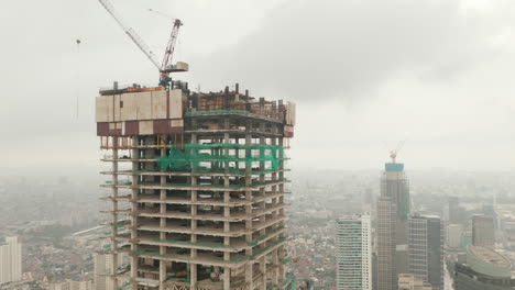 Vista-Aérea-Del-Pedestal-Basculante-De-Un-Alto-Rascacielos-En-Construcción-En-El-Centro-Urbano-De-La-Ciudad-En-Un-Día-Nublado