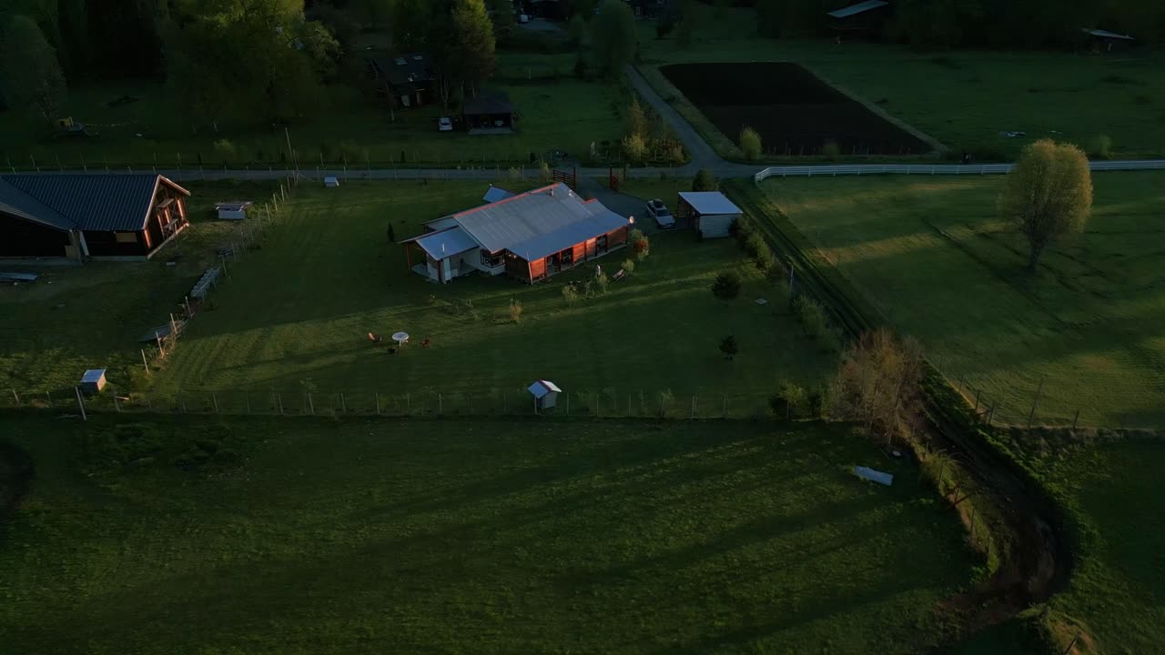 Droneoptagelse over en landejendom og frodige grønne marker i den gyldne time, med spredte landbrugsbygninger omgivet af træer under varmt sollys.