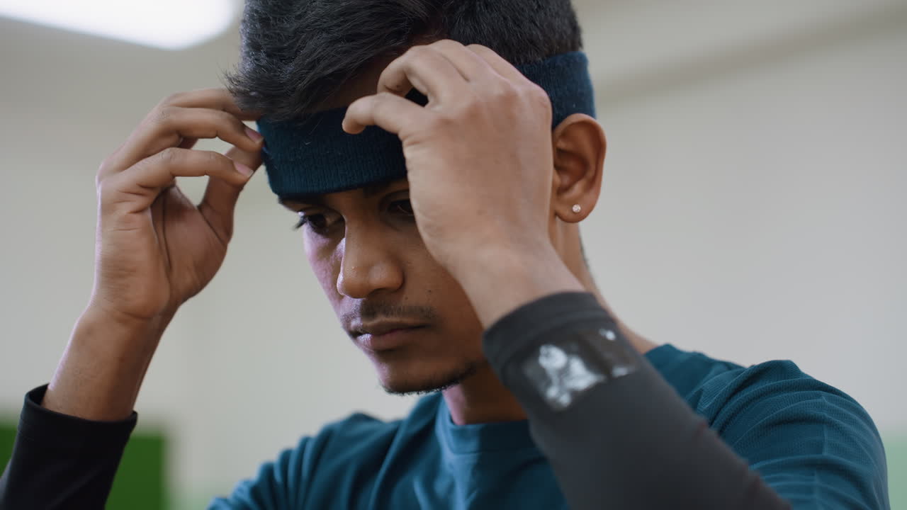 Close up of tennis player adjusting blue headband with both hands, focused expression, fluorescent indoor light shining above with soft blur background in training hall
