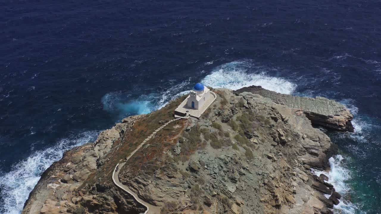 una vista aérea de interés de una pequeña iglesia en una isla griega, entre los acantilados y el mar, con turistas acercándose y de pie