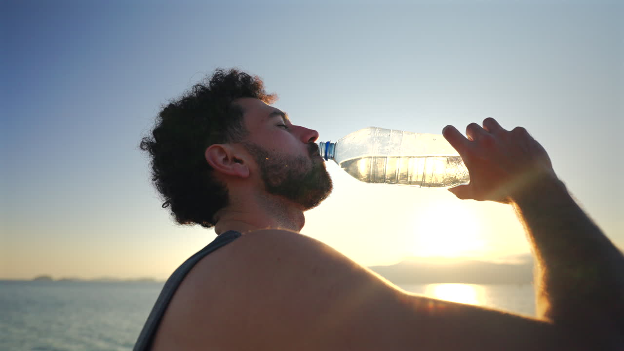 Close-up of a man’s head and shoulders as he drinks water in the sun by the water’s edge