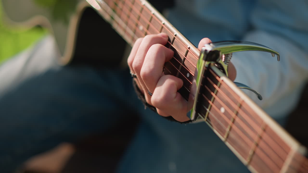 primo piano del cambio del capotasto su una chitarra, sessione di pratica energica che mostra lo spostamento del capotasto e le transizioni di accordi, mani che regolano la tastiera con precisione, musicista in denim che mostra le forme, parco all'aperto