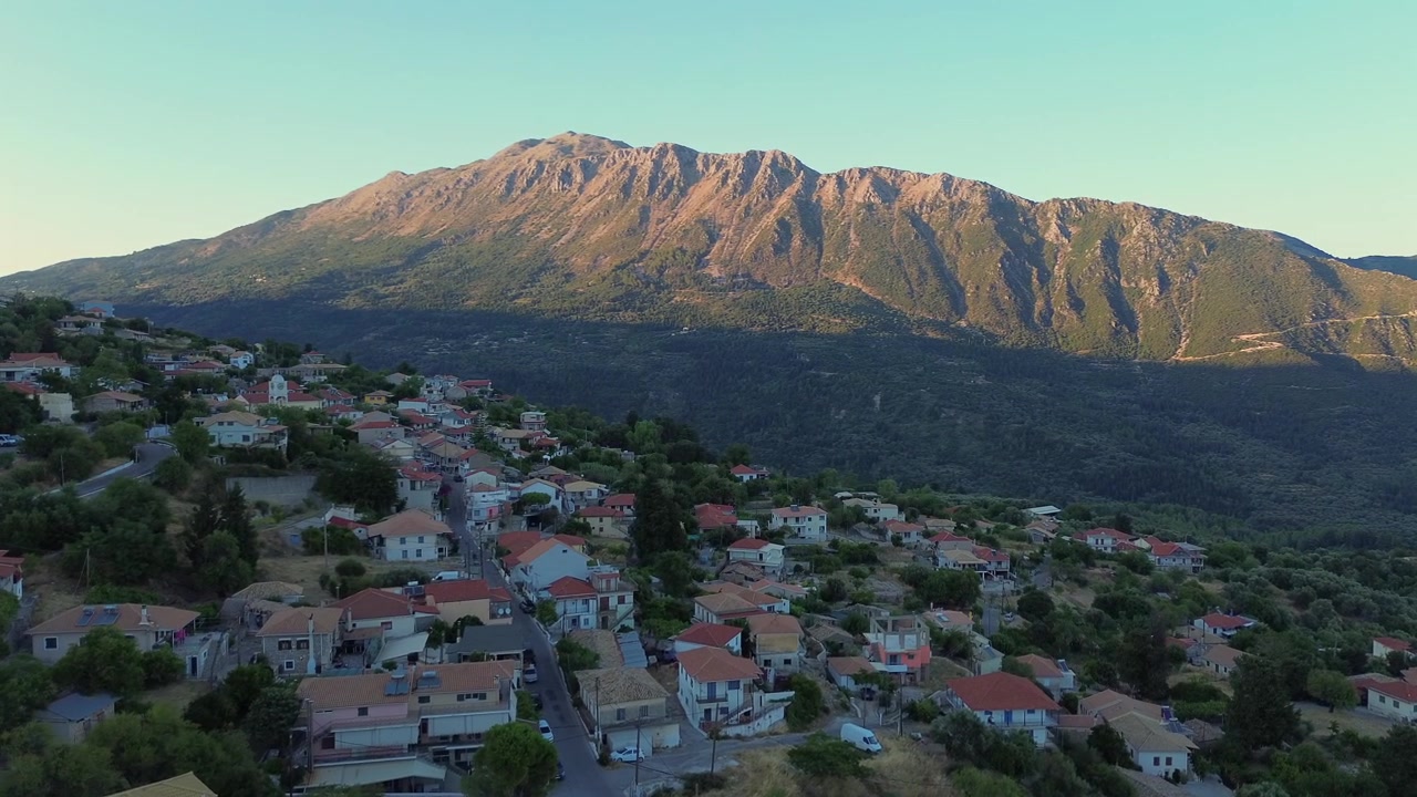 Ampia ripresa panoramica con drone che si sposta a sinistra da una maestosa cresta montuosa per rivelare le case tradizionali del villaggio di Agios Petros a Lefkada, Grecia. Splendido paesaggio rurale di campagna, di giorno