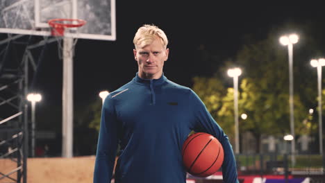 Porträt-Eines-Selbstbewussten-Basketballspielers,-Der-Einen-Ball-Hält-Und-In-Die-Kamera-Blickt,-Während-Er-Nachts-Auf-Einem-Außenplatz-Steht-1