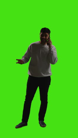 Vertical-Video-Full-Length-Shot-Of-Young-Man-Talking-On-Mobile-Phone-Standing-Against-Green-Screen-With-Low-Key-Lighting-3