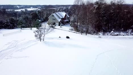 A-drone,-aerial,-birds-eye-view-of-a-child-sitting-on-a-sled-getting-pulled-by-an-atv-4-wheeler-with-another-child-riding-on-the-back