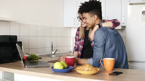 Cute-couple-of-two-young-people-cuddling-and-using-laptop,-surfing-the-web-in-the-kitchen.-Youth,-relationship,-living-together-goals.
