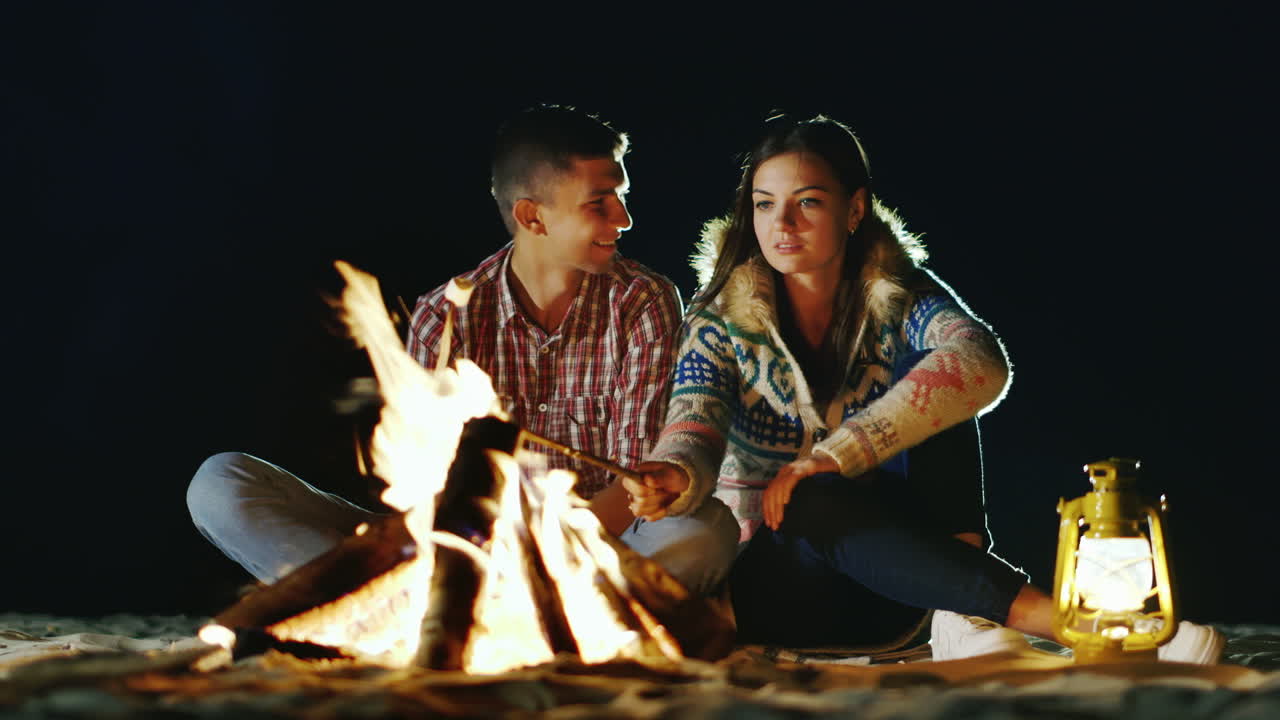 남자와 여자는 모닥불 주위에서 편안한 저녁에 막대기 불에 구운 마시멜로를 굽습니다.