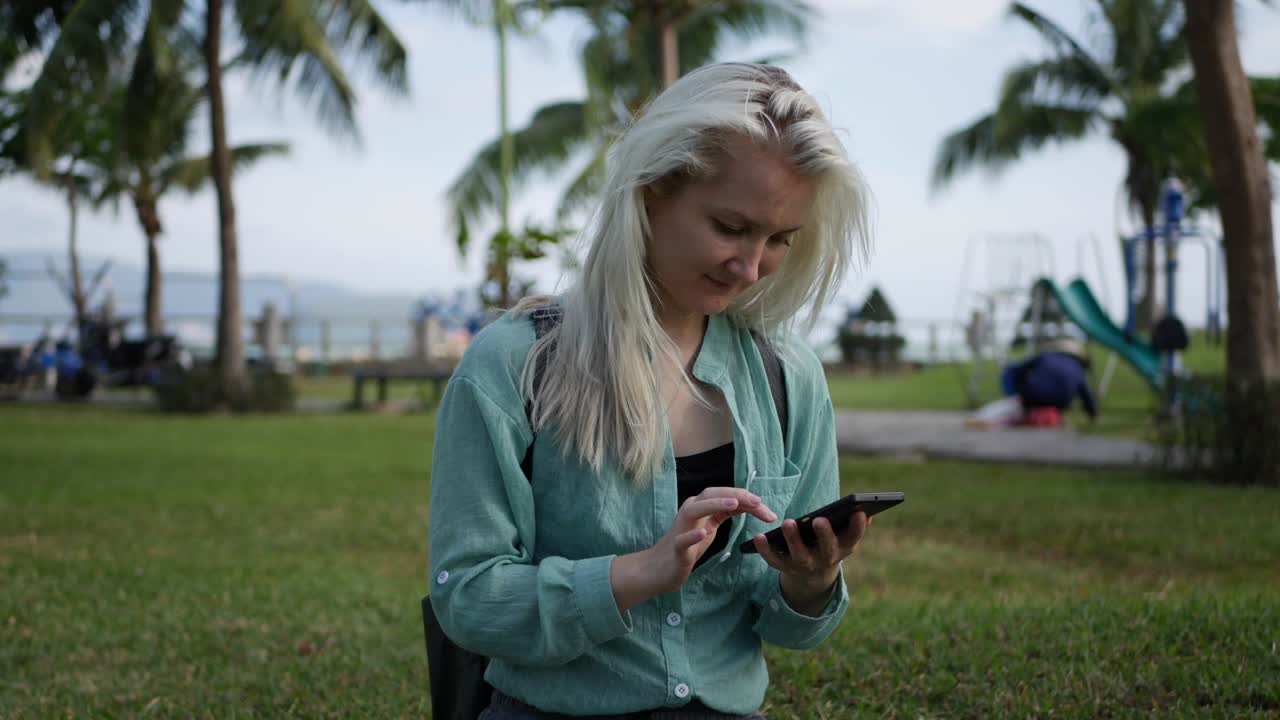 Beautiful slim woman with long blonde hair in green shirt sits on the ground and using smartphone over background the park. Girl on the square touching screen and smile