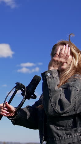 attractive pop singer dances outdoors. beautiful young blonde woman in jeans clothes singing into the microphone on the sky background. vertical video