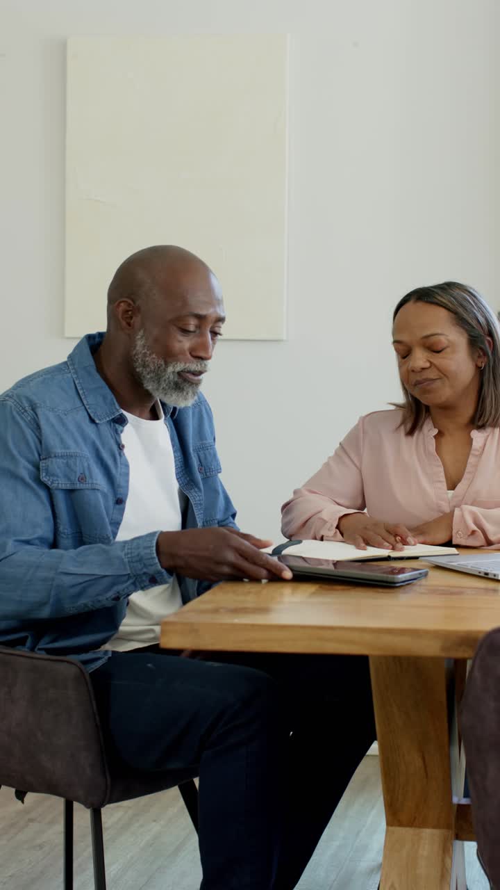 Vertical video: Diverse couple checking bills at home table, man handing tablet while woman writing