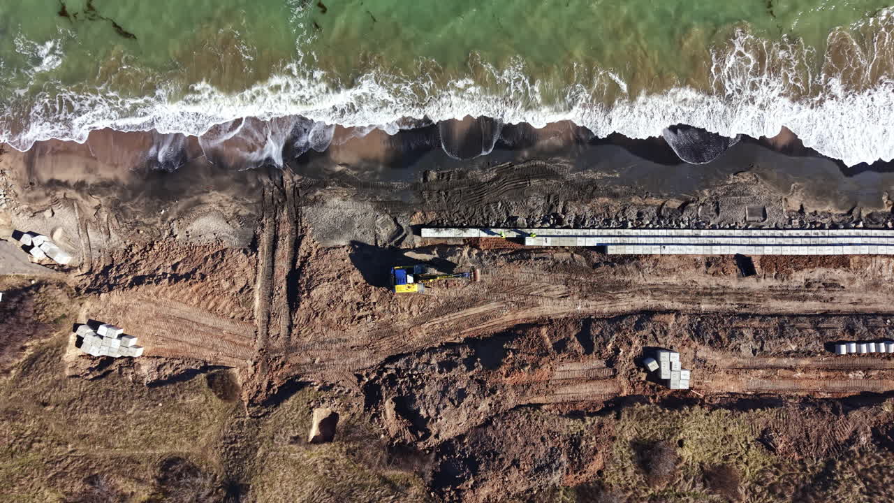 Cantiere edile lungo la costa con mezzi pesanti, in prossimità di uno specchio d'acqua, durante il giorno.