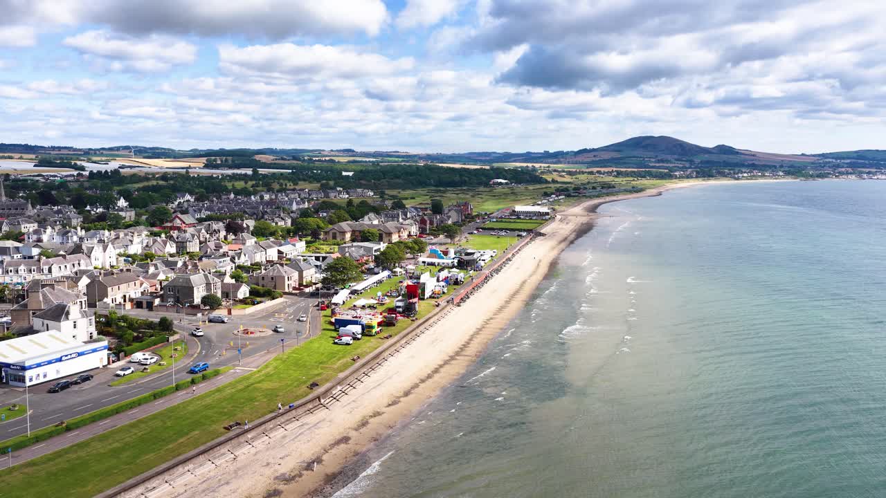 Drone glides above seaside town, sandy beach, and seawall under bright daylight with scattered clouds