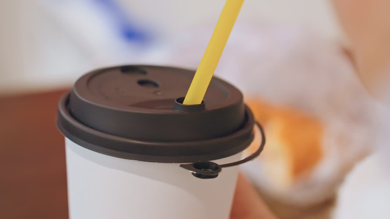 Mom and son straw through lid closeup, mother inserting yellow straw into takeaway cup lid so child can sip warm drink, tight focus on lid and fingers with pastry blurred in background, bright