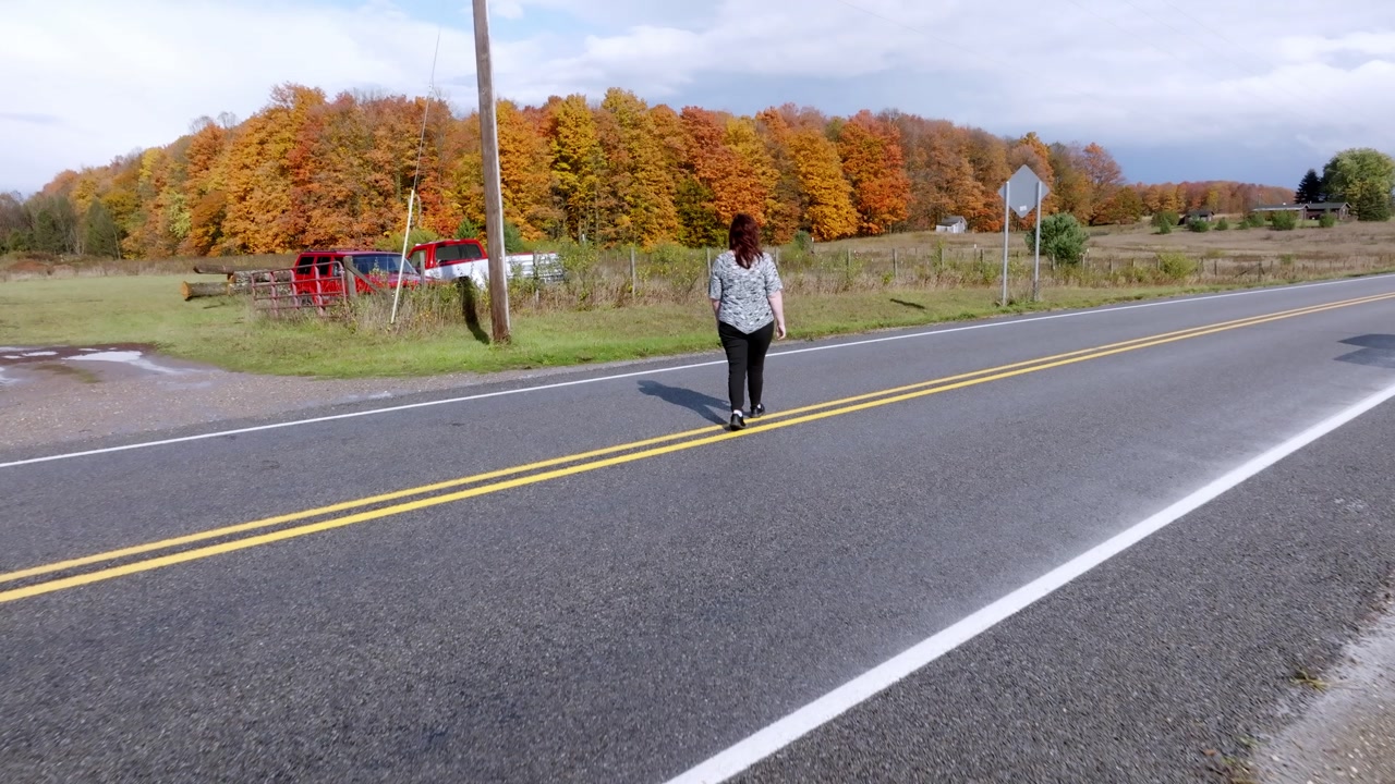 Middle aged caucasian woman walking on rural road in Northern Michigan with fall colors and drone video behind.