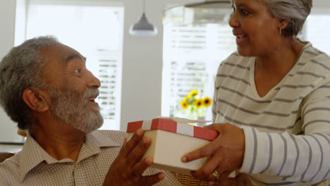 Front-view-of-senior-black-woman-giving-gift-to-senior-man-at-dining-table-in-a-comfortable-home-4k