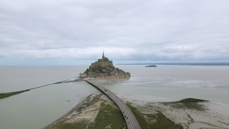 people walking and bus driving along bridge of mont saint michel during low tide on cloudy day, normandy in france