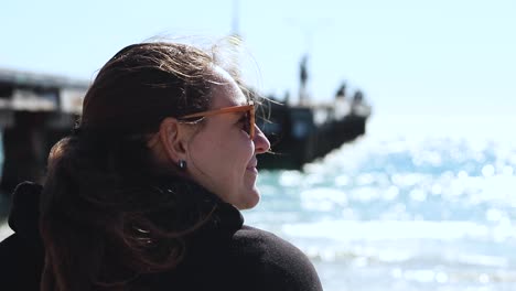 Un-Primer-Plano-De-Una-Mujer-Con-Una-Chaqueta-Negra-Con-Capucha-Y-Gafas-De-Sol-Disfrutando-De-Un-Día-Soleado-Y-Ventoso-En-La-Playa-De-Coogee,-Perth,-Australia-Occidental