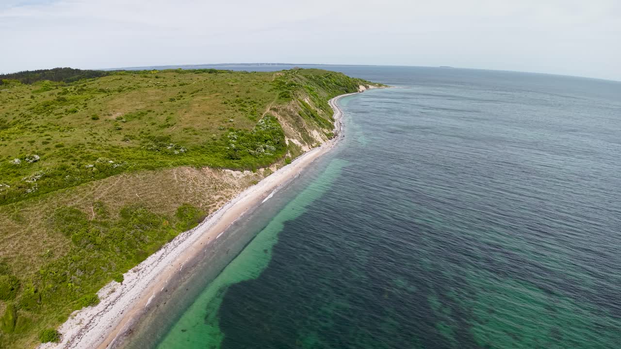 Luftaufnahme mit einer Drohne entlang eines schmalen, weißen Sandstrands mit klarem, türkisfarbenem Wasser und einem grünen, bewaldeten Hang in Dänemark