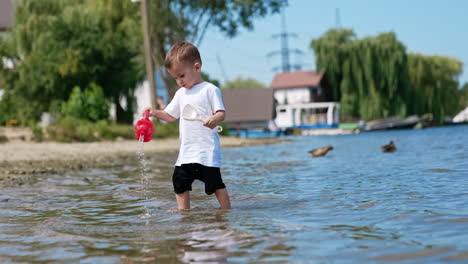 little kid stands in river pouring water from his red watering can. cute caucasian toddler playing on the beach in summer.