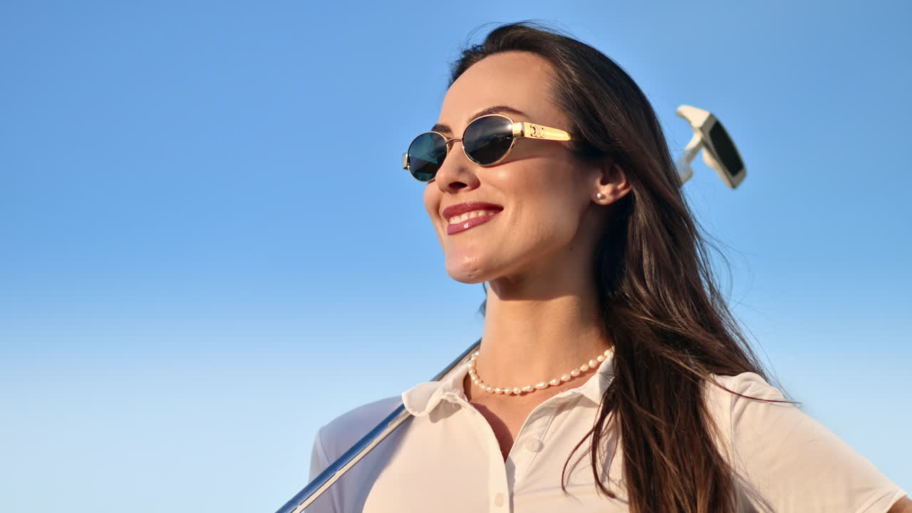 Woman wearing sunglasses, holding a golf club on her shoulder with the blue sky on the background
