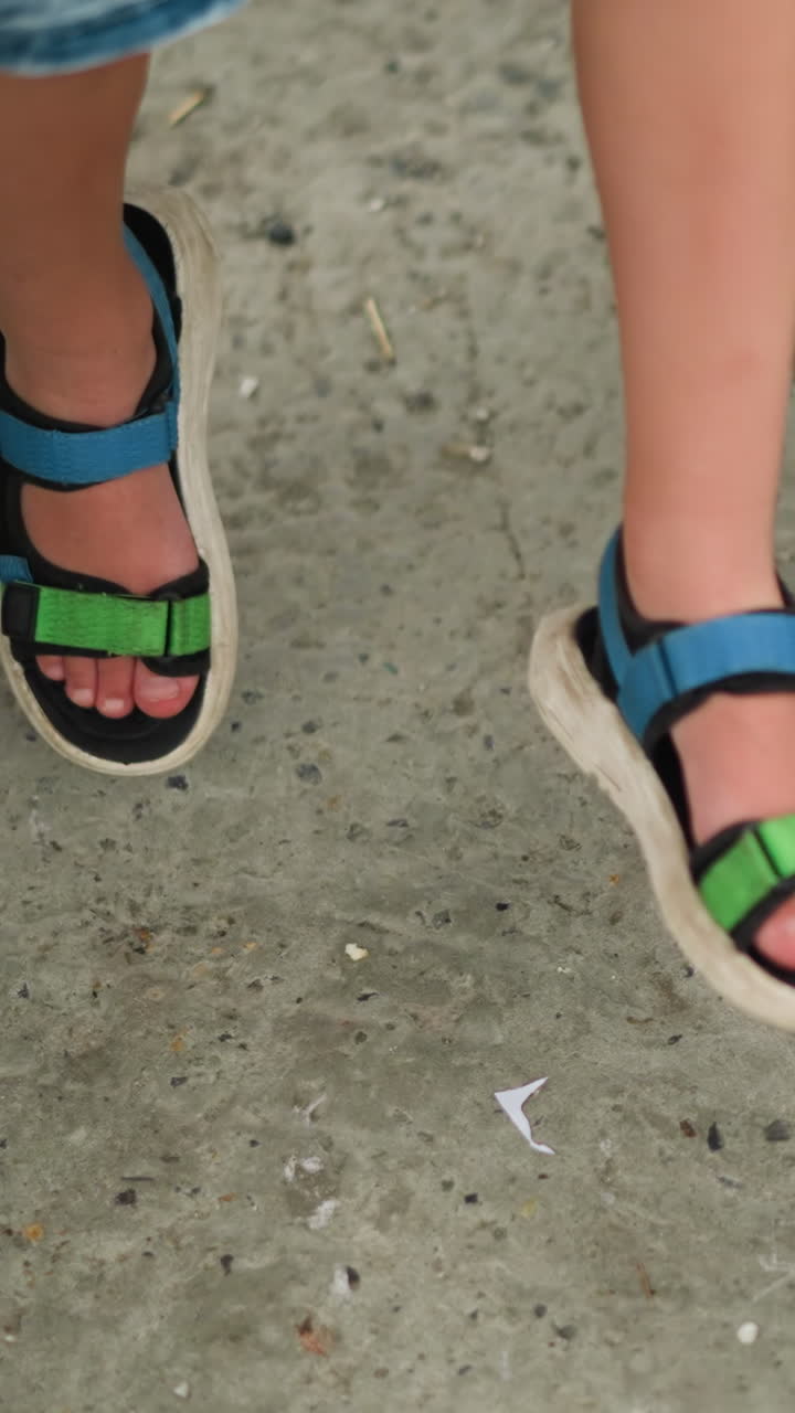 파란색과 초록색 샌달을 입은 아이의 클로즈업, 앉아있는 동안 다리를 흔들고, 녹색 자전거가 근처에 주차되어 있으며, 왼쪽에는 다른 아이의 다리의 부분적인 시각이 있습니다.