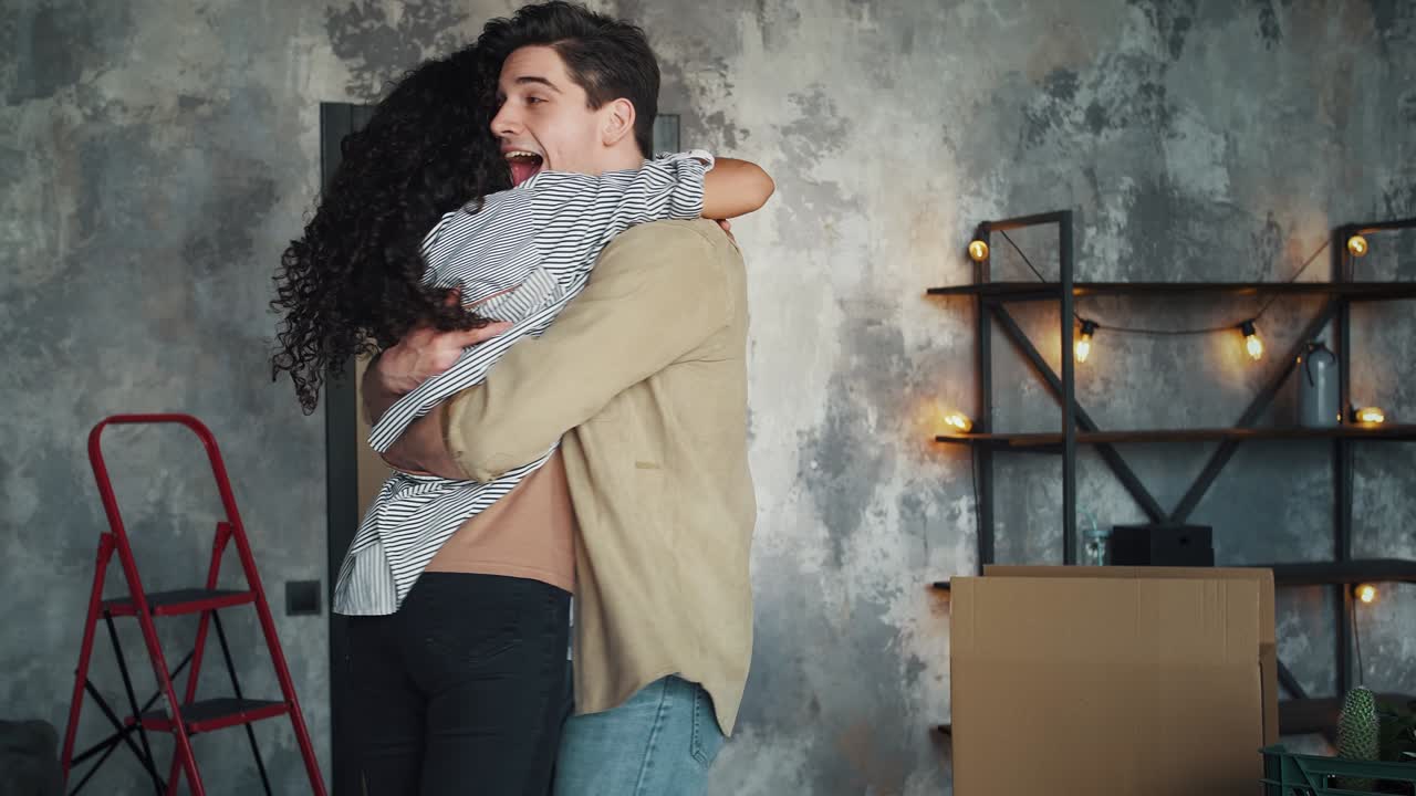 새 아파트로 이사하는 배우자, 남자가 여자를 키우고, 포장 상자와 함께 방에서 그녀를 포옹하고 둘러싸고, 웃고, 기하고, 느린 동작