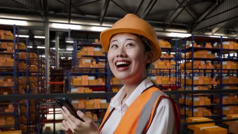 vista lateral de cerca de una ingeniera asiática con casco de seguridad usando un teléfono inteligente mientras está de pie en el almacén con estantes llenos de productos de entrega