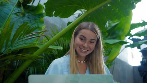 una chica en un vestido azul remotamente en línea trabajando detrás de una computadora portátil y mirando a la pantalla el patio trasero con plantas verdes