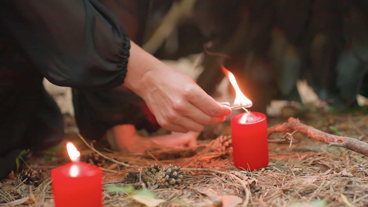 Close up of hand lighting red candle on forest floor surrounded by pinecones, dry leaves, and twigs. Warm flame flickers gently, creating mystical mood of ritual