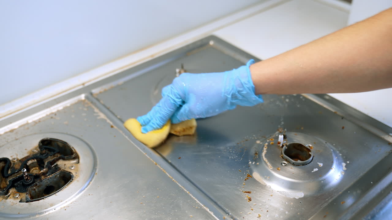 Pulizia profonda della superficie del fornello. Una persona che indossa guanti blu strofina un fornello da cucina sporco con una spugna, rimuovendo efficacemente sporco e residui.