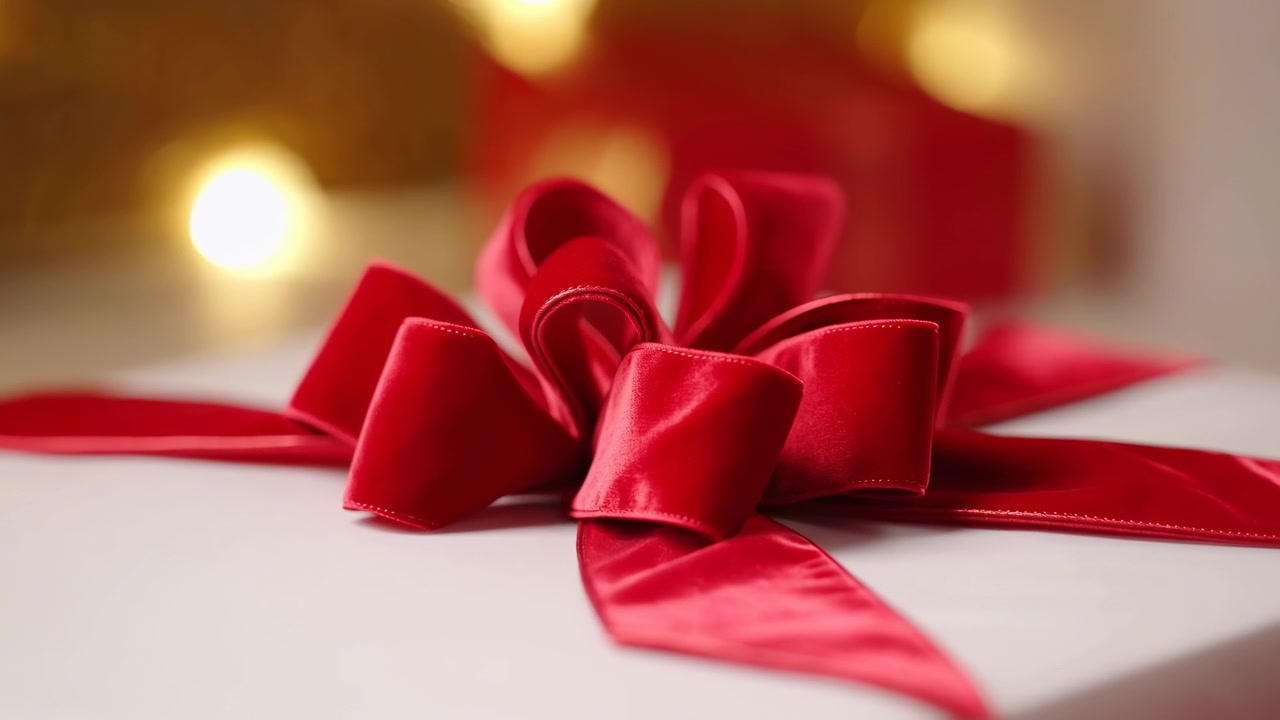 Close-up view of red satin ribbon and bow on smooth white gift box, with warm blurred lights, soft shadows, glossy fabric sheen, diagonal tail, refined holiday packaging mood, with gift, present