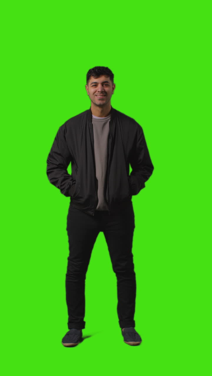 녹색 스크린 앞에 서서 팔을 접는 평범하게 옷을 입은 젊은 남자의 수직 전체 길이의 초상화