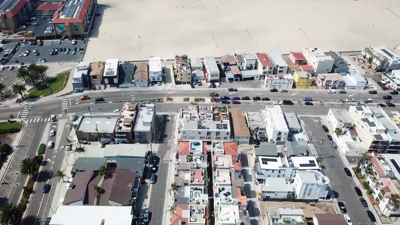 Drone flies above coastal neighborhood in Redondo Beach then tilts up to reveal wide sandy beach and shoreline on a bright day. Clean residential rooftops palm lined streets and light traffic