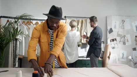 young designer takes measurements and smiles at camera while a man and blonde woman colleagues choose clothes on the background