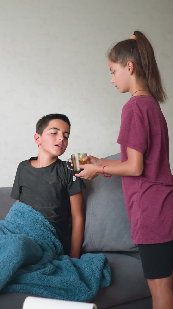 Young girl caring for her sick cousin lying on the couch, handing him a cup of coffee while he gets up to drink it, demonstrating love and support in a home environment