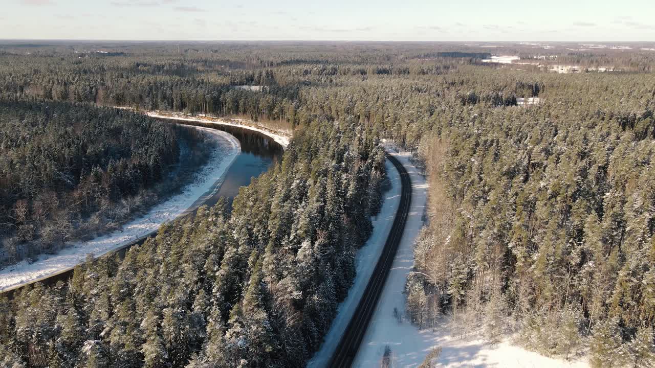 Aerial view of a winding river through a dense snow-covered forest in winter, with a curving road running alongside. A serene and scenic natural landscape.