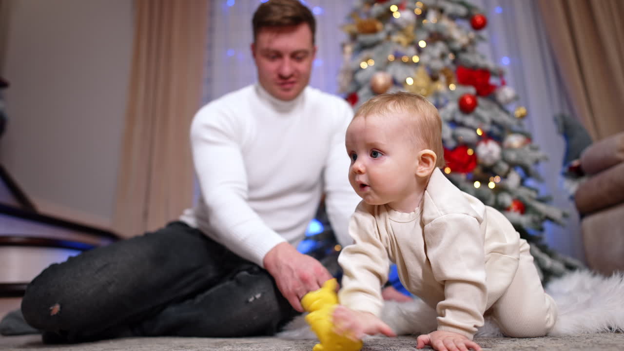 Lovely toddler crawls by the floor heading to something g interesting. Dad sits behind near the Christmas tree watching his son. Low angle view.