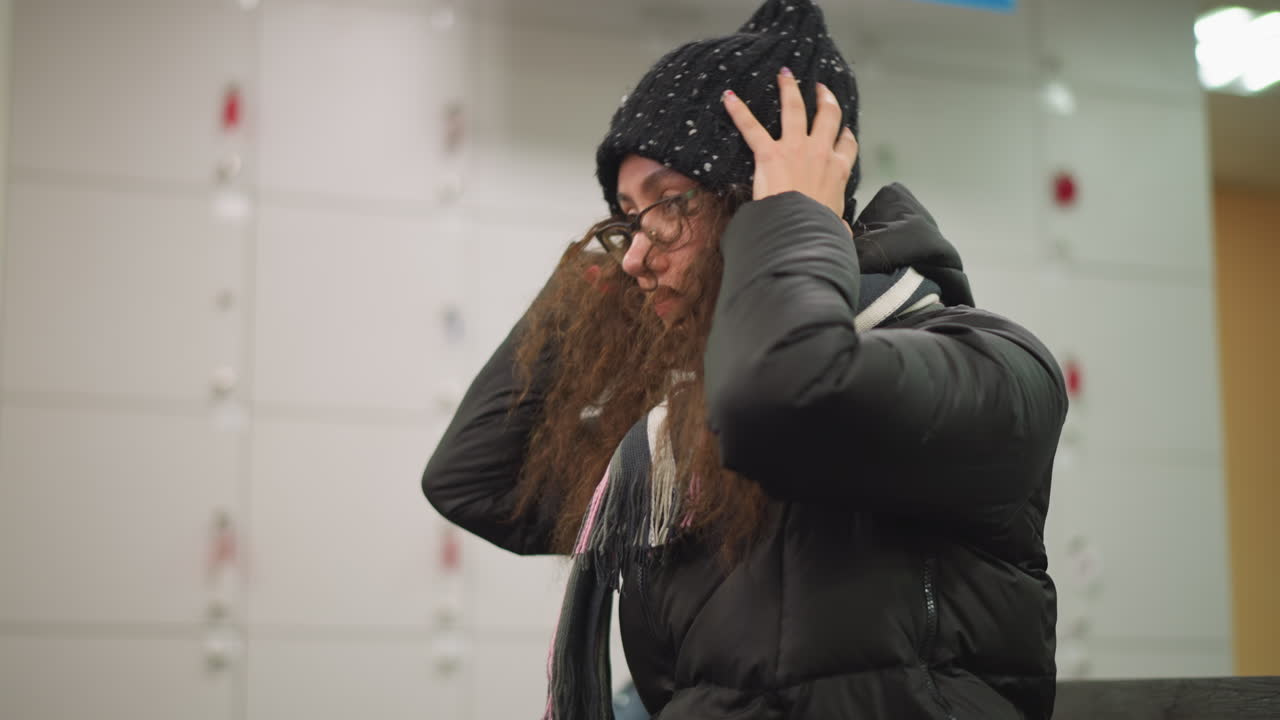 Girl wearing black jacket and striped scarf adjusts messy curly hair with hands indoors, head tilted down in candid natural pose, capturing authentic lifestyle winter moment with casual elegance expression