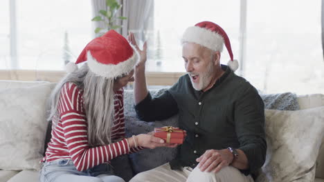 middle aged caucasian couple in santa hats gifting at christmas at home, slow motion