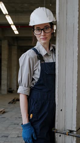 young professional woman builder wearing protective safety gear standing confidently next to concrete pillar within unfinished industrial construction project interior