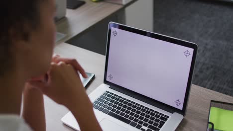 Mixed-race-businesswoman-sitting-at-desk-and-using-laptop-with-copy-space-on-screen-in-office