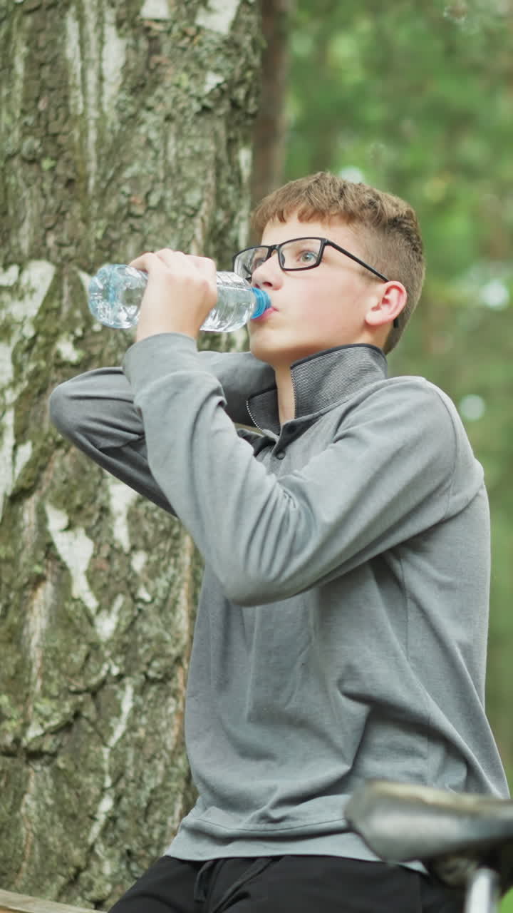 안경을 쓰고 회색 장갑을 입은 어린 소년은 물 병에서 술을 마시며, 높은 초록색 나무로 둘러싸인 숲에서 휴식을 취하고, 자전거가 배경에 그의 에 주차되어 있습니다.