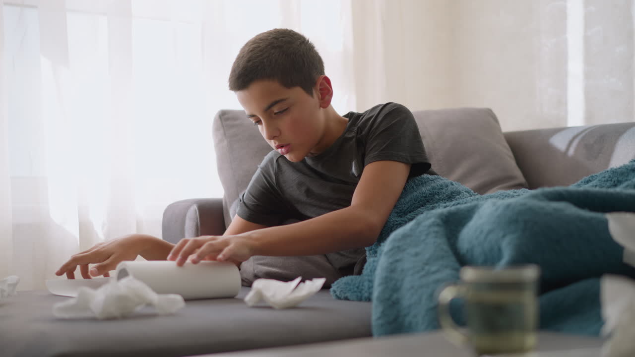 Close-up of glass cup and tissue scattered around with sick boy lying on couch reaching for tissue paper, used tissues around him while recovering from cold or flu at home