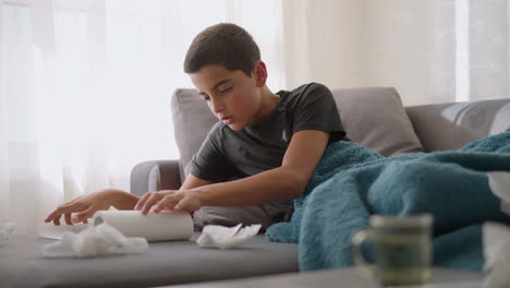 close-up of glass cup and tissue scattered around with sick boy lying on couch reaching for tissue paper, used tissues around him while recovering from cold or flu at home