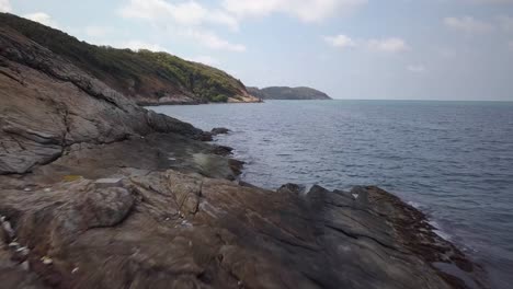 aerial: a forward slow movement and slowly rising the altittude showing the landsacape: tropical island with a rocky shore, steep hills, and a bay, with range of mountains in the distance