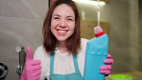 portrait of a joyful and confident brunette cleaning lady in a white t-shirt and a blue apron who points to the detergent in her hand and gives it a like in a pink rubber glove in the bathroom of a modern apartment while cleaning