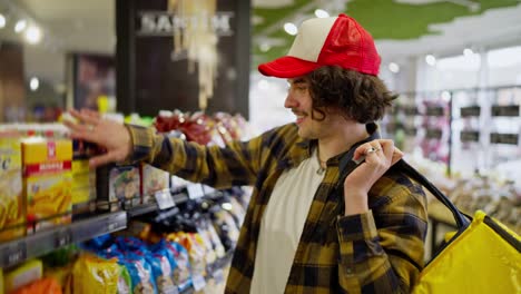 Un-Repartidor-De-Comida-Confiado,-Con-Una-Gorra-Roja-Y-Blanca,-Camina-Cerca-De-Las-Filas-De-Comestibles-Y-Coloca-Los-Productos-Que-Necesita-En-Una-Bolsa-Amarilla-Antes-De-La-Entrega.