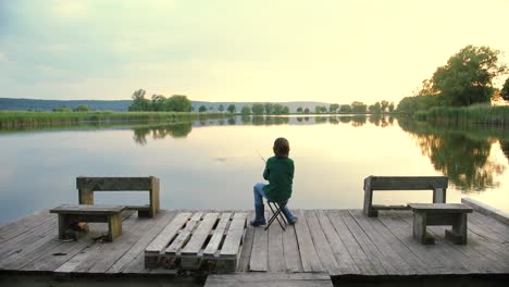 rear view of a teen boy sitting on the lake pier and fishing at sunset