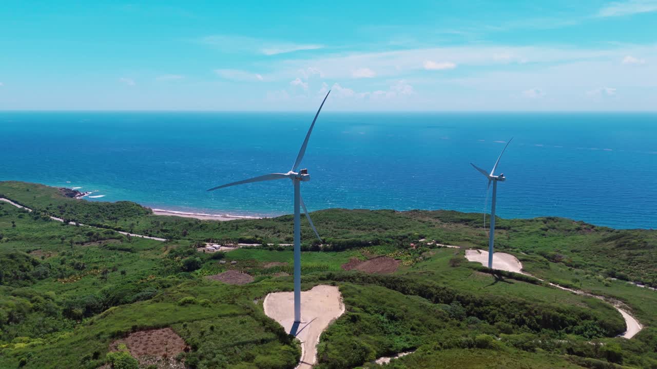 Aerial of two wind turbines spinning on green coastal hill, generating clean energy with blue Caribbean Sea in background, Barahona, Dominican Republic